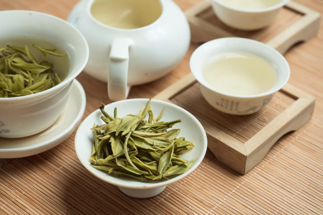 Fresh green tea leaves and brewed tea in white porcelain cups and teapot on a bamboo mat.