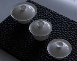 White bone china gaiwans together on a dark textured tea tray